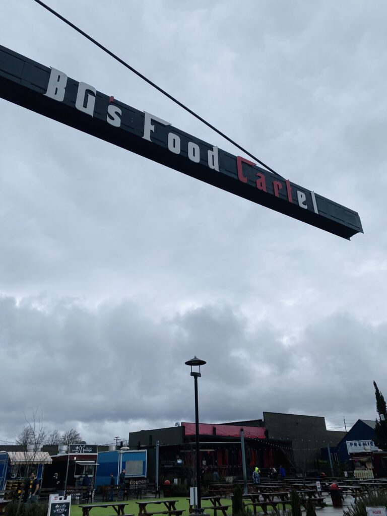 BGs Food Cartel main sign, located in downtown Beaverton, Oregon, with food carts and picnic tables in the background.