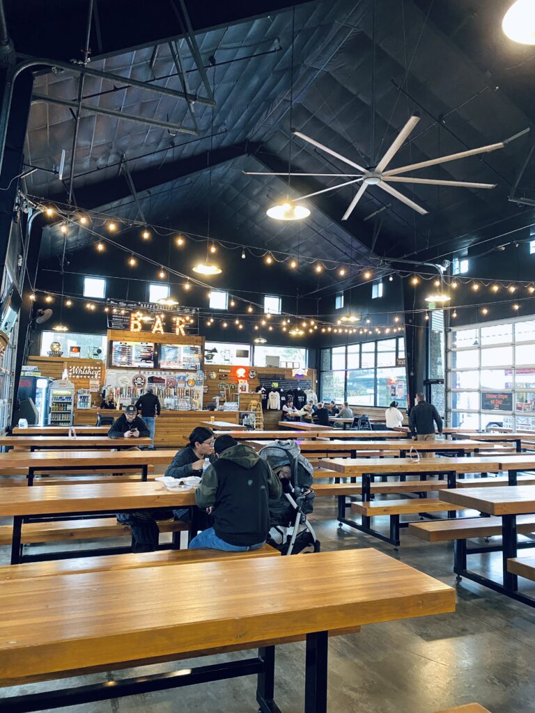 Inside area of the Happy Valley Station Food Truck park. Many tables and people sitting. A bar located in the back with taps.