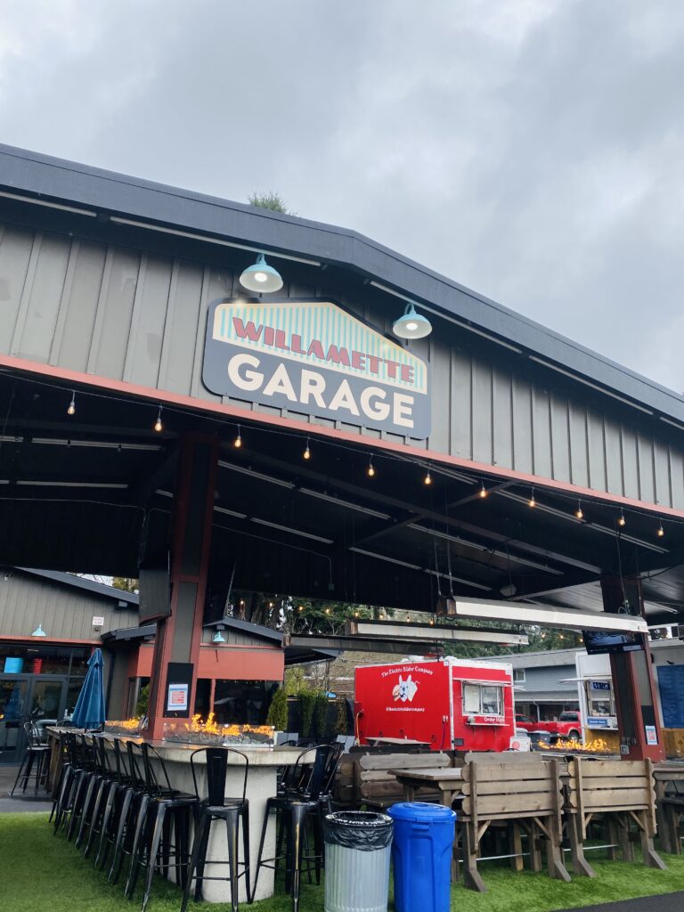 Willamette Garage food cart park, located in West Linn, Oregon. Food trucks in the background with seating area in the front. Seating area has fireplaces on the tables, bar seating and picnic tables.